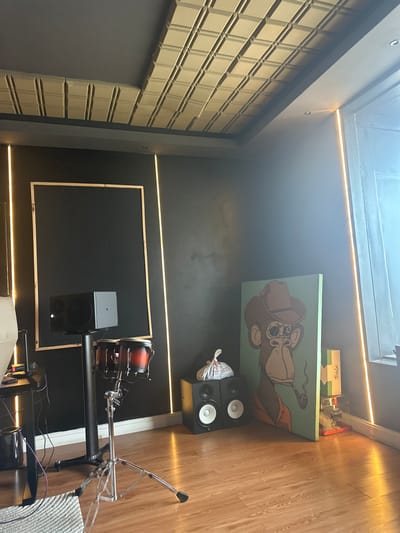 The image shows a music recording studio. There is a wall with soundproofing panels on the left and a wooden floor on the right. A painting of a cartoon dog is mounted on the wall above a pair of studio monitor speakers. A drum set with bongos is in the lower right, and a framed black acoustic panel is on the wall to the lower center. The environment is indoors, with modern lighting. The location appears to be a professional music-related space, although the exact city of Itirin, Nigeria, cannot be confirmed solely from the visual content of this image.