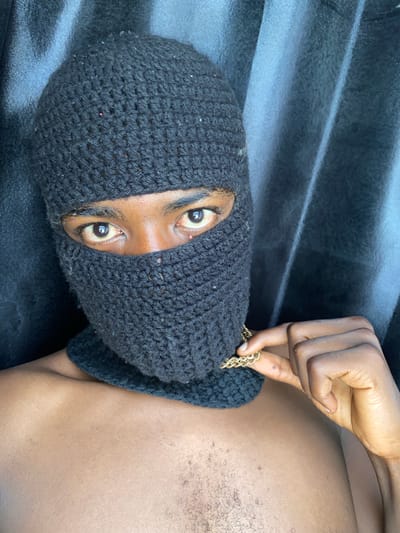 A close-up, high-angle shot features a person wearing a black knitted balaclava, revealing only their eyes. A dark-skinned hand holds a gold chain near the edge of the balaclava, adjacent to the person's torso. The skin tone of the torso matches that of the hand. The person is lying on a textured dark gray or black surface. There is no information about Barakin Akawo, Nigeria, visible in the scene.