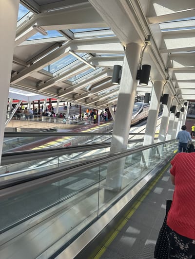 The image depicts an elevated pedestrian walkway in Madrid, Spain, potentially within a transportation hub like a train station or airport. The walkway is constructed of steel and glass, allowing natural light to filter in. Black cylinder-shaped spotlights are mounted on the steel framework. People are visible both on the moving walkway and on a platform below, suggesting a multi-level structure. One or more high-speed trains are visible on the tracks below. Some people are using luggage while moving on the walkway.