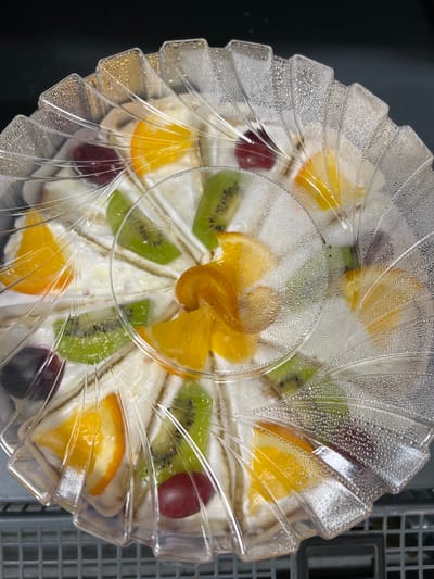 The image shows a close-up, top-down view of a fruit tart housed in a clear plastic container. The tart is cut into multiple triangular slices, each decorated with pieces of fruit and cream. Visible fruits include slices of orange and kiwi, as well as whole grapes. The plastic lid of the container is covered in condensation, suggesting it may have been refrigerated. The tart is arranged on a surface that is partially visible, with a dark background on the left and a grey, gridded surface on the right, possibly indicating a refrigerator shelf or a kitchen counter. The overall impression is of a freshly prepared dessert ready for consumption. No people are visible in the image. The lighting suggests an indoor setting. There is no visible text in the image. The location context of Sinyavino, Russia, does not provide any visual cues within the image itself.