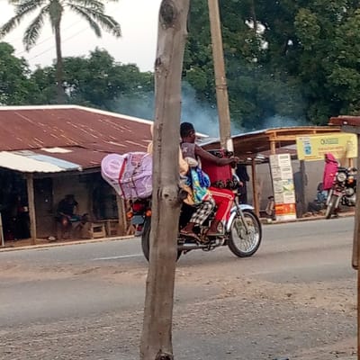 The image depicts a daytime street scene in Mararaba, Nigeria, under what appears to be an overcast sky or hazy conditions, with some smoke visible in the distance. The central subject is a person riding a red and silver motorcycle, moving from left to right on a paved road. The rider, wearing a dark top and plaid trousers, has large bundles wrapped in purple and other fabrics secured to the back of the motorcycle.

A prominent tree trunk stands in the foreground, partially obscuring the view. To the left of the road, there are open-fronted buildings with corrugated metal roofs, one of which contains a person sitting. To the right, similar roadside structures are visible, including a stall or shop. A yellow sign hanging from one of these structures clearly displays the words "OUTDOOR" and "BUSINESS". Another person, dressed in a pink garment, stands near a parked motorcycle further down the road. The background is filled with lush green foliage and a tall palm tree, indicative of a tropical environment. The road surface appears worn.