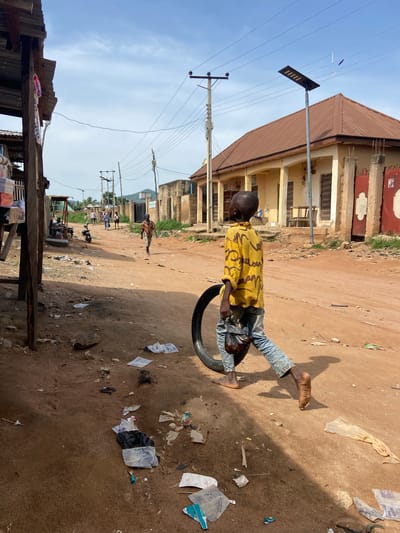 A young boy, appearing to be of African descent, is captured in a still image, energetically pushing a tire along a dusty, unpaved road. He is dressed in a yellow t-shirt with a black graphic print and blue jeans, his bare feet contrasting with the textured denim. The boy's posture suggests movement, as if he's in the midst of play or engaging in a task that requires effort.

The scene unfolds in a somewhat impoverished rural or semi-urban setting, likely Kuson, Nigeria, given the context. The road is characterized by its dry, reddish-brown earth, scattered with debris and trash, including plastic bags and paper scraps. Behind the boy, a building with a brown, corrugated metal roof stands prominently. Its facade is weathered, with visible windows and architectural details that suggest a modest, possibly older structure. In the background, more buildings, utility poles with overhead wires, and sparse vegetation can be seen. The sky above is a clear blue with a few wisps of clouds, indicating a bright, likely daytime scene. The lighting suggests it could be late morning or early afternoon, with the sun casting distinct shadows on the ground, including the boy's own shadow. The overall atmosphere is one of everyday life in a developing area, where children find ways to entertain themselves with simple objects. There is no visible text in the image.