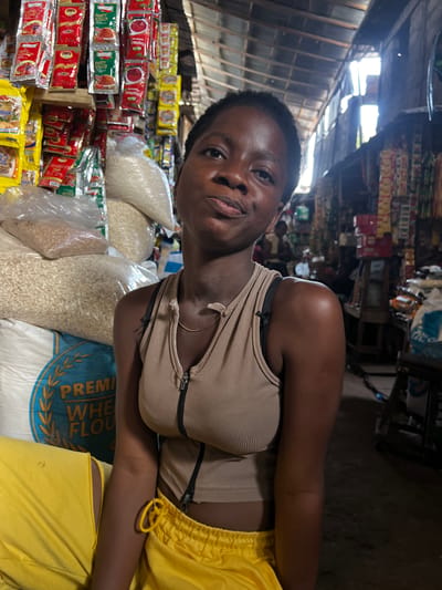 In Nkpor, Nigeria, a young Black woman is photographed from the chest up, seated in what appears to be a market stall. She has short, dark hair and is looking directly at the camera with a neutral expression. She is wearing a beige, ribbed tank top with a zipper down the front and yellow drawstring shorts. Behind her, the market is stocked with various goods. To her left, large sacks of what appears to be grain or flour are stacked high, with one sack clearly labeled "PREMIUM WHEAT FLOUR" in blue and yellow lettering. To her right, shelves are filled with packaged food items, including brightly colored sachets of spices and sauces. The market setting is rustic, with a corrugated metal roof visible overhead and natural light filtering in from the open sides. In the background, other market-goers are visible, suggesting a bustling commercial environment.