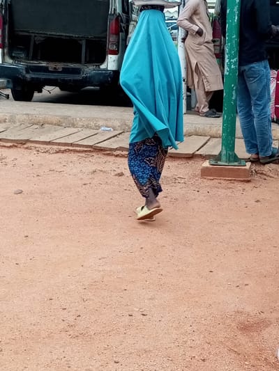 The image depicts a scene on a dirt road in the city of Gombe, Nigeria. In the foreground, a person is walking away from the camera, wearing a vibrant turquoise hijab that drapes over their body and a patterned blue and white skirt. They are also wearing light-colored sandals. To the left of the person, the rear of a white van is visible, appearing to be parked. The van's doors are open, revealing a dark interior. To the right, a green metal pole stands on a concrete base, and beyond that, two other individuals are standing. One person, on the far right, is wearing blue jeans and dark sandals, with their back to the camera. Next to them, a person in a light beige robe with their hands in their pockets, also facing away from the camera, is leaning against a surface. The ground is a dusty, reddish-brown earth mixed with small pebbles. Wooden planks are laid out on the side of the road.