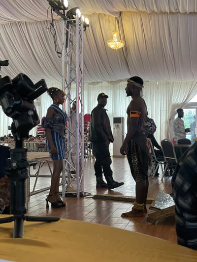 This media file captures an indoor event in Barakin Lerawi, Nigeria, likely a celebration or performance. The scene is set within a draped tent, suggested by the abundance of white fabric creating a soft, diffused background. A prominent crystal chandelier hangs from the ceiling, casting a warm glow and indicating an evening or dimly lit indoor setting.
The primary subjects are several individuals, seemingly engaged in a presentation or performance. In the foreground, a person in traditional attire, adorned with a patterned skirt and beaded anklets, stands looking upwards. Behind them, a person in a black leather jacket and cap is also present, standing stoically. Further back, another individual, shirtless and wearing a headband and an orange armband, is positioned looking down. In the background, a person in a striped shirt is seated, looking at a phone.
The event appears to involve some form of performance or ritual, given the formal attire of some individuals and the stage-like setup with lighting rigs and truss structures. The lighting suggests it's an indoor event, possibly in the evening. The floor is tiled with a warm wooden pattern. The overall mood is one of anticipation or engagement with the ongoing activity. No visible text is present in the media file.