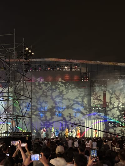 The image captures an outdoor fashion show at night in Chongqing High-tech Zone, China. A large stage structure with lighting and display panels is the backdrop, featuring an abstract tree-branch pattern projected onto the surface. A line of people are observing the show, with many holding up their phones to record or photograph the event. Some stage scaffolding is visible in the foreground.