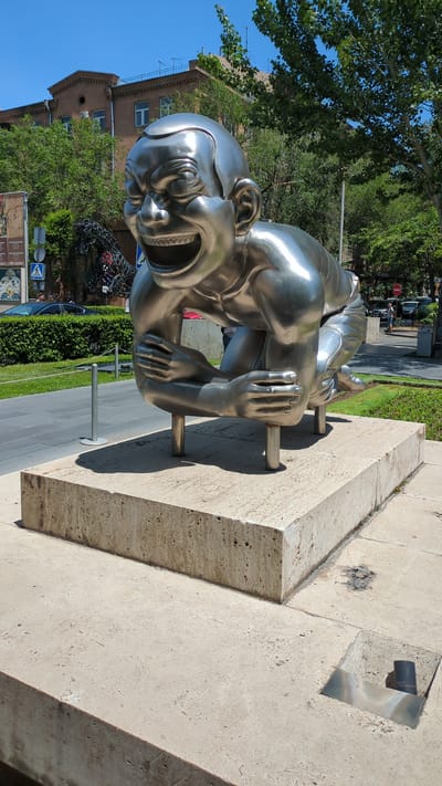 This image presents a highly reflective, polished metallic sculpture of a stylized human figure in a crouched, laughing pose, situated outdoors under a clear blue sky. The sculpture, crafted from a silver-toned metal, depicts a figure with an exaggeratedly wide-open mouth revealing teeth, squinted eyes, and deep facial wrinkles indicative of intense laughter. Its arms are bent, with hands clasped or intertwined over its knees. The figure is supported by four cylindrical metal posts that connect its underside to a light-colored, rectangular stone plinth made of porous travertine.

The plinth rests on a broader expanse of similar light-colored stone paving. In the lower right foreground, a square recessed opening in the paving contains a small, dark cylindrical object. To the left of the sculpture, a grey sidewalk with embedded metal bollards runs parallel to a neatly trimmed green hedge. Beyond the hedge, a street is visible with parked vehicles, including a black sedan, and a pedestrian crossing sign. In the mid-ground, a dark, abstract metal sculpture or structure composed of numerous interconnected elements is partially visible behind the main subject.

To the right, additional green foliage and trees line a street in the distance, where a red stop sign and other vehicles are discernable. A person is partially visible near the stop sign. The background features a multi-story brown building with a distinct roofline, multiple windows, and several rooftop antennas, positioned beneath a bright, cloudless blue sky. The scene is illuminated by direct sunlight, producing strong reflections on the sculpture and sharp shadows. This sculpture, known as "Men" (or "Happy Men"), is located within the Cascade Complex in Yerevan, Armenia.