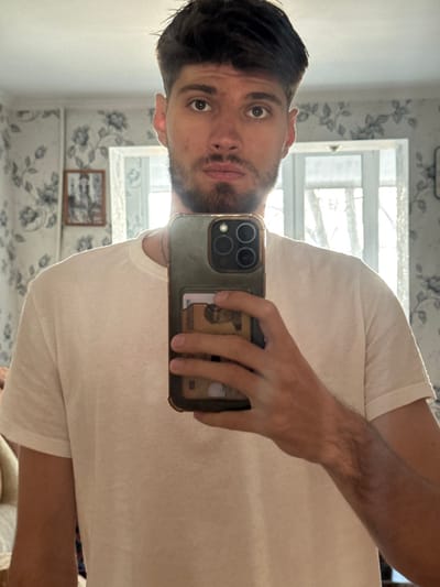 A male individual is depicted from the upper chest upward, reflected in a mirror. He possesses short, dark brown hair and a short beard and mustache. His expression is neutral, and he wears a plain white short-sleeved t-shirt with a thin chain around his neck. He is holding a dark-colored smartphone, equipped with a multi-lens camera array and a rugged case, in his right hand to capture the reflection. The phone case features a card slot containing several cards, including one with a visible gold-toned circular element. The background shows a wall covered with light-colored floral patterned wallpaper. To the right of the subject, a large window provides bright ambient light, revealing faint outlines of trees outdoors. To the left, a small, framed picture is mounted on the wall. The ceiling and a white decorative molding are visible at the top of the frame. The scene is illuminated by natural light entering through the window. This indoor setting is located in the city of Ciorescu, Moldova.
