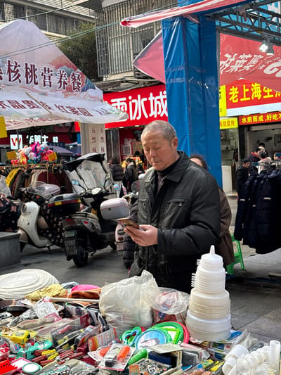 An outdoor street scene in Jiangjin, China, depicts a bustling market environment. In the immediate foreground, a market stall displays a variety of consumer goods, including stacks of white circular plastic items, a clear plastic bag filled with merchandise, packaged tools such as screwdrivers and tape measures, various plastic toys, and a stack of nested white plastic funnels.

A middle-aged to elderly Asian male, with short, greying hair and wearing a dark, possibly black, zipped-up jacket over a dark high-necked garment, stands in the mid-ground. He is focused on a smartphone, which he holds with both hands and looks down at the screen. Another individual is partially visible behind him.

The background features several parked motor scooters or electric bicycles. Beyond these vehicles, multi-story urban buildings with numerous storefronts are visible. Overhead, white and red awnings or tarps extend across the street. Prominent signs with Chinese characters include a red sign for "蜜雪冰城" (Mixue Bingcheng), a yellow sign for "老上海生煎" (Lao Shanghai Shengjian), and a large white awning bearing "核桃营养餐" (Walnut Nutrition Meal). To the right, clothing racks display various garments, and a blue vertical support pole is visible. The ground is paved, and the lighting suggests an overcast or diffused daylight condition.