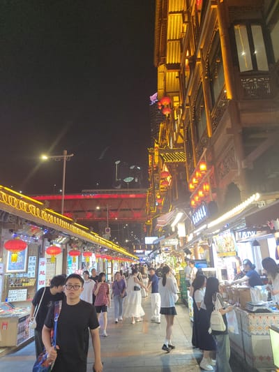 This is a nighttime, eye-level shot of a bustling pedestrian street in Yuzhong District, China. The street is lined with traditional wooden buildings illuminated by warm yellow lights and red lanterns, creating a vibrant, festive atmosphere. Several people are walking along the cobblestone street, some browsing at stalls. On the left side of the image, there are stalls selling various goods, with posters and signs in Chinese. The right side features more stalls and a row of shops with awnings. In the background, a large, multi-level bridge with red and yellow lighting spans across the sky, with modern skyscrapers visible beyond it. The overall scene depicts a lively commercial and entertainment area at night.