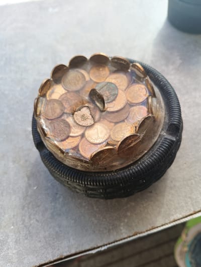 A close-up, high-angle shot shows a homemade ashtray, likely located outdoors on a light gray, slightly textured surface, possibly a patio or table. The ashtray is constructed from a black rubber tire inner tube cut into a circular shape, forming the base. Encased in clear resin on top of the tire is a collection of copper-colored coins, arranged in a circular pattern. Some of the coins are partially submerged in the resin, while others are fully covered. A larger, darker coin or token is placed prominently in the center of the resin-filled area. The overall impression is of a rustic, repurposed item. The image does not contain any identifiable individuals or specific city landmarks that would indicate a location in Karnobat, Bulgaria.