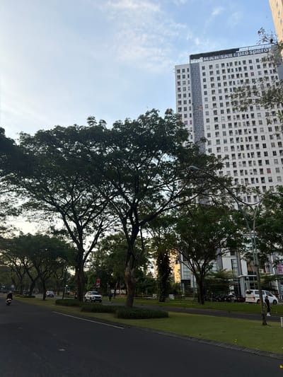 In Entalsewu, Indonesia, a large apartment building, "MANSAR PROSPERO," stands tall against a partially cloudy sky. Its facade is a grid of numerous windows, with a decorative cloverleaf pattern on the left side. In the foreground, a paved road runs alongside a well-maintained green median strip with trees. Several vehicles, including cars and motorcycles, are visible on the road and parked in the background. People are seen walking and riding motorcycles, suggesting a lively urban environment. The scene is set in a city, with the presence of modern architecture and infrastructure.