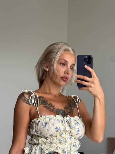 A person with blonde hair and visible tattoos on their chest and shoulder takes a selfie in a room with a light grey wall. They are holding a dark blue smartphone in their right hand, which is positioned in front of their face. The person is wearing a white and blue floral print top with tie shoulder straps. The background is plain, indicating an indoor setting, likely a bedroom or dressing area. The lighting appears natural, coming from the side. The overall scene is casual and personal. There is no indication of the specific location within Kuta Selatan, Indonesia, but the environment suggests a typical residential or hotel room.