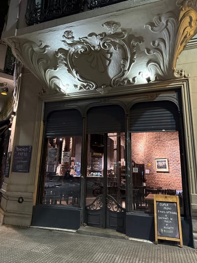 The image captures the illuminated entrance of a bar or restaurant at night, set on a tiled city sidewalk. The building features elaborate, light-colored decorative stucco or stone carvings on the left side of the doorway, illuminated by a small spotlight, showcasing flowing and organic architectural motifs.
The entrance comprises large, dark-framed glass doors, providing a view into the establishment's interior. Inside, the space features an exposed brick wall, with a framed picture hanging on it. Tables and chairs are dimly visible, along with shelves stocked with bottles or bar supplies, suggesting a cozy, inviting atmosphere. The overall lighting within the venue is warm.
To the right of the entrance, a freestanding chalkboard sign, framed in wood, advertises a "SUPER PROMO" (Super Promotion). The text, written in white, details the offer: "1 PINTA ARTESANAL" (1 Craft Pint) "+ BASTONES DE MUZZA" (Mozzarella Sticks) "e NUGGETS" (and Nuggets) for "$11.000".
Below the chalkboard, a dark blue sign is affixed to the wall, stating "TENEMOS PATIO" (We Have a Patio), "Climatizado" (Air-conditioned), and "Apto fumadores" (Smoking permitted). An Instagram handle "@imaginerestaurant" is also present on this sign. Other visible text elements inside the bar include signs for "CHIMPANCE", "TU MAMA TAMBIEN", and various beer types or brands such as "BEERDIER", "IPA", "SCOTTISH", "KOLSCH", and "HONEY". The name "LUIS" is partially visible. The sidewalk is paved with textured tiles. No people are directly visible, but the open establishment suggests a vibrant urban scene.