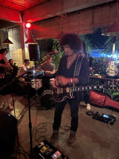 The image depicts an indoor performance space, possibly a bar or restaurant, in Kuta Selatan, Indonesia. Red lighting illuminates the ceiling, while bamboo blinds cover windows or walls. A band is performing, with a lead guitarist in the foreground playing a dark-colored electric guitar and wearing a black open-neck shirt and dark pants. He has a large, curly hairstyle and a necklace. To his left, a drummer is seated behind a drum kit, wearing a gray t-shirt. Other band members and audience members are visible in the background. Audio equipment, including microphones, speakers, and a pedalboard with multiple effect pedals, are arranged on the floor. A red guitar case is visible on the right side of the frame. The overall atmosphere suggests a live music event in a casual setting.