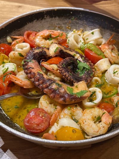 The image displays a close-up, overhead view of a seafood dish served in a rustic, shallow pan. The dish prominently features a large piece of cooked octopus tentacle as the centerpiece, surrounded by shrimp, calamari rings, cherry tomatoes (both red and yellow), and green peppers. The seafood is swimming in a vibrant, golden-yellow broth, glistening with oil and flecked with chopped parsley. A metal spoon is visible in the lower left portion of the frame, partially submerged in the broth, suggesting the dish is ready to be eaten. The pan itself appears to be made of metal with a slightly textured, dark interior and a rim that shows some wear. The dish is placed on a wooden surface, likely a table, with a visible wood grain. The lighting suggests an indoor setting, possibly a restaurant. The specific location of Tel-Aviv, Israel is not indicated by any visual cues within the image itself.