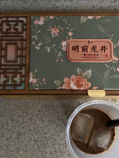 This image captures an overhead view of a decorative wooden box with a floral pattern and a plastic cup filled with a beverage and ice cubes. The box appears to be made of wood with intricate lattice work on the left side and a textured, green fabric covering on the right, adorned with pink and orange floral designs. A copper-colored plaque with Chinese characters and English text "YICHA YIYI YI REN SHENG" is affixed to the fabric. The cup, positioned at the bottom right of the frame, contains a brown liquid with two large ice cubes and a black straw. The background is a gray, speckled surface, resembling a countertop or floor. The scene is set on a concrete surface, suggesting an outdoor or patio setting, potentially in a city park or courtyard in Querétaro, Mexico. There are no people visible in the frame, and no specific actions or interactions are depicted, beyond the placement of the objects. The lighting is natural, casting soft shadows.