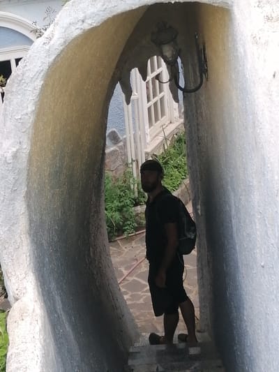A man stands within a peculiar, organically shaped archway, the rough, textured surface of which dominates the foreground. The archway appears to be made of concrete or a similar material, with a light, yellowish interior and a rougher, white exterior. A traditional-style outdoor lantern hangs from the ceiling of the arch. Beyond the arch, a building with light blue walls and a white-trimmed window is visible. Lush green foliage grows around the building and on the ground. The ground is paved with irregular stone tiles. The man, wearing a dark t-shirt, dark shorts, a dark beanie, and sandals, is seen from the waist down and is looking over his shoulder towards the camera. He has a backpack on. The lighting suggests it is daytime, likely late afternoon due to the warm tones and shadows. The overall scene evokes an unusual architectural space, possibly a garden or an eccentric building entrance, set in a warm climate given the vegetation. Da Lat, Vietnam is a possible location context.