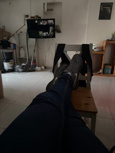 A first-person perspective image shows a person's legs clad in dark denim jeans, with gray Nike sneakers resting on a light wooden chair. The scene is indoors, with light-colored tiled flooring. In the background, a television is mounted on a white wall, along with shelving units. Other items include a vacuum cleaner, white containers, and miscellaneous objects on the shelves. An abstract painting is visible on the wall above the television. It is impossible to determine whether or not the location is in Jerusalem, Israel.