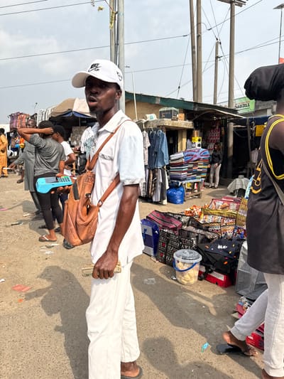 A man wearing a white shirt, white pants, a white baseball cap, and a brown leather shoulder bag stands in a crowded outdoor market. He holds what appears to be a stack of money in his left hand. In the background, vendors display various goods, including clothes, fabrics, and packaged food. Wires hang overhead, and other people are walking through the market.