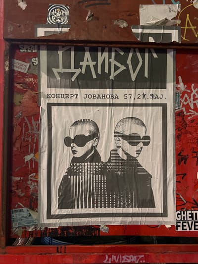 The image shows a poster adhered to a weathered, red metal surface, possibly a bulletin board or a door, in an urban outdoor environment. The poster itself is largely monochrome, featuring two stylized, bald figures wearing large, dark sunglasses, one slightly behind the other. Their faces are rendered in a graphic, almost pixelated style. Above the figures, in bold, stylized Cyrillic lettering, appears the word "ДАНБОГ". Below the figures, in smaller Cyrillic text, it reads "КОНЦЕРТ ЈОВАНОВА 57,2К. МАЈ". The poster is creased and slightly torn in places, suggesting it has been exposed to the elements.

Surrounding the central poster, other elements are visible. To the left, there are fragments of other posters and stickers, some showing peeling red paint underneath. One visible sticker has the word "ARTMORIA" and a QR code. To the upper left of the main poster, a circular sticker with an eye-like graphic is present. To the right of the main poster, red paint is heavily graffitied with black spray-painted lines and lettering. Two small stickers are visible on this red surface below the main poster, one with "GHETTI" and another with "FEVE". At the very bottom of the frame, a red metal bar is visible, with some graffiti in white marker.

The overall impression is that of urban street art and advertising, common in public spaces in Belgrade, Serbia, where Cyrillic script is used. The style of the poster and the surrounding graffiti suggest a specific artistic or cultural context.