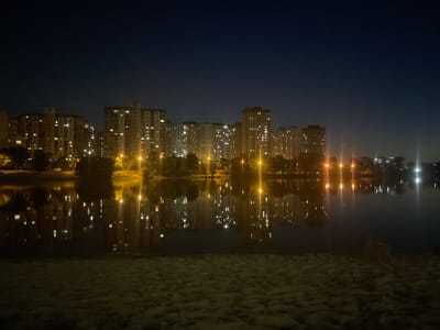 This media file depicts a nighttime cityscape in Kyiv, Ukraine, with a focus on residential buildings lining a body of water. Tall apartment blocks, illuminated by numerous lit windows, dominate the background, their reflections shimmering on the dark surface of the water. Streetlights cast warm yellow glows, creating streaks of light that are also mirrored in the water. The foreground consists of a sandy or earthy embankment, with indistinct textures suggesting it might be a beach or riverbank. In the far right of the frame, a small, light-colored object, possibly an animal or a rock, is partially visible on the embankment. The overall mood is tranquil and slightly melancholic, characterized by the stillness of the night and the soft glow of artificial lights against the dark sky. The time of day is clearly night, and no specific weather conditions are evident beyond the calm water surface. There is no visible text in the image.