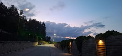 The image depicts an outdoor scene at dusk or twilight in Bania, Bulgaria. A paved road or street curves gently uphill and to the right, extending into the background. On the left side, a low stone retaining wall runs parallel to the road, topped by a dark metal fence. Behind this fence, a dense forest of dark conifer trees slopes upwards, illuminated by several green decorative lights interspersed within the foliage. Multiple streetlights with bright yellow-white illumination are positioned along the left side of the road, casting light onto the asphalt. A dark-colored car is parked facing away from the viewer on the left shoulder of the road. Further down the road, two indistinct figures are visible in the distance.
On the right side of the road, a series of dark wooden fence panels, possibly forming a retaining wall or property boundary, are covered with dark green climbing vegetation. Recessed warm-toned lights are integrated into several sections of this wooden fence, casting a soft, yellowish glow. The fence line extends into the background, where distant mountains are silhouetted against a sky displaying a gradient of blue, purple, and pink hues, indicative of sunset. Overcast conditions are present, with dark clouds prominent across the mid-sky, and lighter, scattered clouds higher up. The overall scene is illuminated primarily by artificial lighting against the fading natural light of evening.