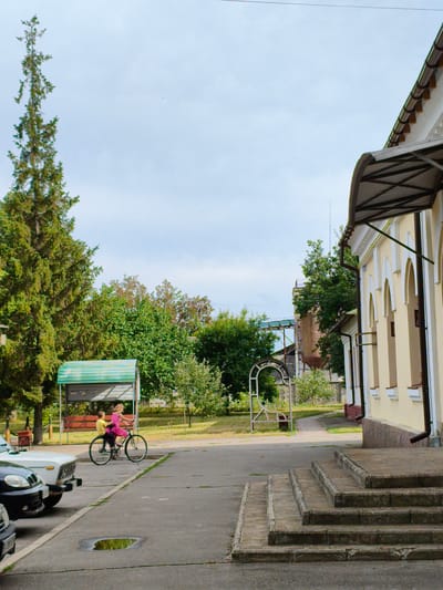 This is a photograph taken outdoors on a cloudy day. The scene depicts a paved area in front of a building with a yellow facade and arched windows. A set of concrete steps leads up to the building's entrance, which is partially covered by an awning. To the left of the building, there are trees and a covered picnic area with benches. 

In the foreground, a boy and a girl are riding a bicycle. The boy is pedaling, and the girl is seated on the back of the bicycle. They are both dressed in casual clothing. The girl is wearing a pink dress. The bicycle appears to be an older model with a large front wheel. 

Several cars are parked on the paved area. One car in the foreground is white, and another is black. A puddle of water reflects the sky on the pavement near the cars. In the background, there are more trees and a distant industrial structure. The overall impression is of a quiet, possibly rural, setting. The sky is overcast, suggesting a day without bright sunshine.