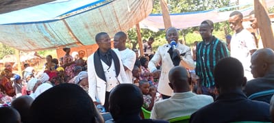 The image depicts an outdoor gathering, possibly a ceremony or event, in what is likely a rural setting near the city of Kalemie, Democratic Republic of the Congo. A large crowd of people, including many women in colorful clothing and head wraps, are seated under a makeshift canopy of blue tarpaulin. Several men, some in formal attire such as white suits, stand in the foreground and appear to be addressing the crowd with a microphone. The environment includes trees and modest structures, suggesting a village or community setting. The overall atmosphere suggests a significant occasion taking place within the community.