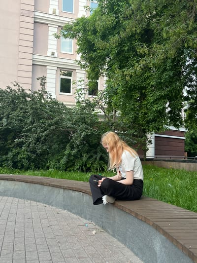 A young woman with blonde hair sits cross-legged on a curved wooden bench in a park setting, looking down at her phone. She is wearing a white t-shirt and black pants. The bench is situated in front of a grey stone retaining wall, with a paved area in the foreground. Behind the bench, a lush green lawn is visible, with a large tree providing shade. In the background, a pale pink building with white trim and windows stands adjacent to a dark brown wooden structure. The scene appears to be a public outdoor space in the city of Krasnoyarsk, Russia, possibly a park or a courtyard.