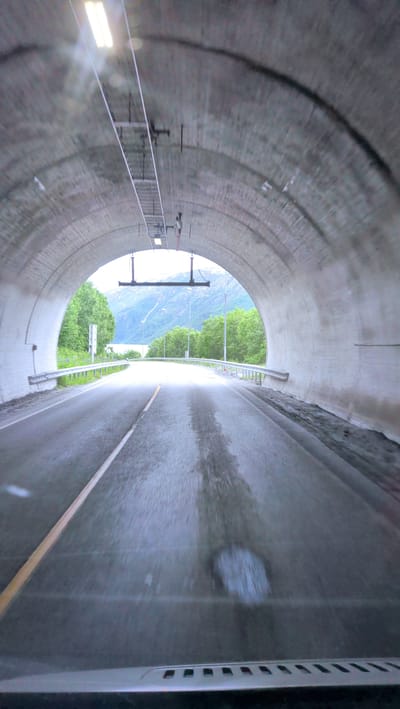 This media file captures the view from inside a vehicle as it drives through a tunnel towards daylight. The tunnel is constructed from rough, grey concrete with an arched ceiling. Fluorescent lights are visible on the ceiling, casting a bright glow. A metal railing lines the right side of the road inside the tunnel.
As the vehicle exits the tunnel, a scenic view of a fjord is revealed. Lush green trees line the sides of the road, and the water of the fjord stretches out towards distant, hazy mountains. The sky appears bright, suggesting it is daytime. The road surface inside the tunnel shows signs of wetness, with some darker patches indicating moisture, and a light yellow line marking the lane division. The overall atmosphere conveyed is one of transition, moving from the enclosed darkness of the tunnel to the open, natural beauty outside. There is no visible text in the image. The location cues, such as the fjord and mountainous landscape, combined with the tunnel setting, are consistent with driving in Norway, specifically near Mosjøen.