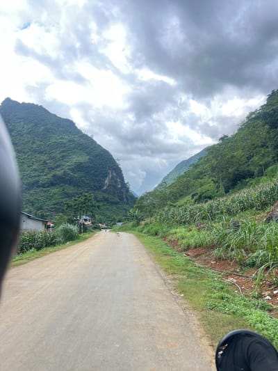 The image depicts a view of a rural road in Cán Tỷ Commune, Vietnam, characterized by steep, verdant mountains on either side. The road itself is unpaved, appearing to be a dirt or gravel track, curving gently into the distance. On the right side of the road, a field of corn stalks grows abundantly, interspersed with taller grasses and some scattered debris. The left side of the road features a few scattered buildings, including what appears to be a simple house with a white exterior, and a small agricultural field. A traffic sign is visible near the buildings. The sky above is overcast with dramatic, dark grey clouds, suggesting impending rain or a storm. The perspective suggests the photo was taken from a moving vehicle, possibly a motorcycle, given the partial view of a black helmet in the foreground. A solitary, small figure, presumably a person, is visible walking on the road in the distance, adding a sense of scale to the vast mountainous landscape. The overall impression is one of a remote, agricultural community nestled within a dramatic, natural environment.