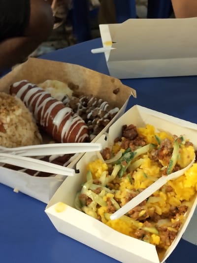 The image shows a close-up view of two takeout food containers filled with food, likely at an outdoor market or street food stall in Davao City, Philippines. The containers are made of white paperboard, and the food appears to be a savory rice dish with meat and vegetables, and a large sausage drizzled with white sauce. Plastic forks are placed on top of the food, suggesting that it is ready to be eaten. The background is blurred, but it appears to be a blue surface, possibly a table or a tablecloth. The lighting suggests it is daytime, and the scene is informal.