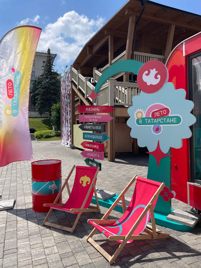 The image depicts an outdoor scene in Kazan, Russia, likely at an event or festival given the festive decorations. In the foreground, two red deck chairs with a light blue pattern are set up on a paved area. To the left of the chairs, a bright red barrel with a light blue and white design stands. Behind the barrel and chairs, a tall, colorful banner displays the text "ЛЕТО В ТАТАРСТАНЕ" (Summer in Tatarstan) and a sun graphic.

A prominent signpost with directional arrows points to various cities: "КАЗАНЬ" (Kazan), "НАБЕРЕЖНЫЕ ЧЕЛНЫ" (Naberezhnye Chelny), "АЛЬМЕТЬЕВСК" (Almetyevsk), "ЗЕЛЕНОДОЛЬСК" (Zelenodolsk), "НИЖНЕКАМСК" (Nizhnekamsk), "АЛАБУГА" (Alabuga), and "МАМАДЫШ" (Mamadysh). A large, light blue, star-shaped sign with the same "ЛЕТО В ТАТАРСТАНЕ" text and a sun graphic is positioned to the right of the signpost. A red, curved structure, possibly part of a food truck or stand, is visible to the far right.

In the background, a multi-story wooden building with a balcony and stairs is visible, suggesting it might be a pavilion or an entertainment venue. Behind the building, a tall evergreen tree stands next to a building with a white facade. The sky is blue with scattered white clouds. The overall atmosphere suggests a sunny day, likely during the summer months. There are no people clearly visible in the image.