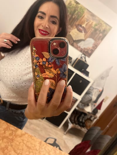 A mirror selfie is captured indoors in the city of Konz, Germany, depicting a female subject and the surrounding room. The woman, smiling, has dark hair, prominent eyebrows, and wears red lipstick. She is dressed in a white eyelet-patterned top, dark blue jeans, and a dark belt. A light-colored ring adorns her left ring finger. Her fingernails are manicured with a light, neutral polish. She holds a dark red smartphone, featuring a multi-lens camera system and a clear protective case decorated with glitter, autumn foliage (berries, leaves), and the reversed "CASETiFY" brand name, in front of her. Her left hand is raised towards her hair.
The reflected background reveals a light-colored wall. A tilted painting on the wall depicts a large pair of lips, possibly holding a cigarette, against a blurred landscape with trees and flowers. Below the painting, a black and gold handbag rests on a white cubby shelf unit containing black storage boxes. To the right of the shelves, an ornate table with dark legs is visible, followed by a dark wooden furniture piece. The floor appears to be light-colored wood. In the foreground of the reflection, a light brown and tan patterned surface (likely a countertop or table) is visible, with a dark, irregularly shaped object on it. Red and grey fabrics are partially visible in the lower right portion of the reflection.