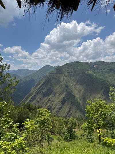 The image shows a view from Lũng Cú Commune, Vietnam, featuring a steep, green, mountainous landscape under a partially cloudy blue sky. The perspective is from within a structure with a thatched roof visible in the left corner. Lush vegetation fills the right side of the frame. A faint path or road is visible tracing along the face of the mountain. The lighting suggests a sunny day. A building can be seen near the top of the mountain.