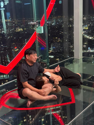 Two individuals are embracing on a glass floor overlooking a city at night. The person on the left is a man wearing a black t-shirt and black shorts, with his arm around the person on the right. The person on the right is a woman wearing a white top and black pants, with her head resting on the man's shoulder and her legs wrapped around his torso. They appear to be in a romantic or intimate embrace.
The setting is a high vantage point, likely a skyscraper or observation deck, with a clear glass floor providing a view of the city below. The city is illuminated by numerous lights, indicating it is nighttime. Red neon lights are visible on the structure of the observation deck, adding a modern aesthetic to the scene. The background shows a dense urban landscape with many buildings and lights stretching into the distance.
The mood seems intimate and romantic, with the couple enjoying a moment together against the backdrop of the vibrant city. The lighting suggests it is evening or night. There is no visible weather information.
The text "VIDEO SCOPE" is visible on a sign in the bottom left corner of the image.
The location context provided is Hanoi, Vietnam.