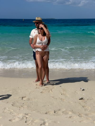 Two individuals, an adult male and an adult female, stand embraced on a light-colored sandy beach in Zanzibar, Tanzania. The male, positioned behind the female, has a beard, wears sunglasses, a straw fedora, a white short-sleeved patterned shirt, and dark shorts. His arms encircle the female's waist. The female, facing slightly left, wears sunglasses, a white crocheted-style spaghetti strap top, and light brown bikini bottoms; her right hand is raised to her forehead. Both subjects are barefoot, standing at the edge of the incoming tide where small white waves break. The ocean exhibits turquoise hues near the shore transitioning to darker blue further out. A yellow buoy is visible in the mid-ground water, with a distant landmass on the horizon under a mostly clear blue sky with sparse white clouds. Strong shadows of the subjects are cast on the sand to their right. The sandy foreground contains visible footprints and small white organic debris.