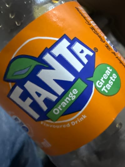 A close-up, angled shot captures the label of a Fanta Orange soft drink bottle. The vibrant orange label dominates the center of the frame, with the bottle itself showing some condensation or water droplets, suggesting it is cold.
The main subject is the Fanta logo, prominently displayed in white block letters outlined in blue, against a blue background. A green leaf design is integrated into the logo's 'A'. Above the main logo, a green speech bubble contains the white text "Great Taste", written upside down relative to the main Fanta logo. To the right of the logo, a green rectangular banner also displays the word "Orange" in white, also oriented upside down.
Other visible text on the orange label includes "Product of The Coca-Cola Company" along the top edge, and "Flavoured Drink" along the right edge, both printed in a dark, likely black, script. The overall impression is a brightly colored, refreshing beverage. There are no people visible, and the background is blurred and dark, offering no specific location cues beyond the product itself. The image appears to be taken indoors.