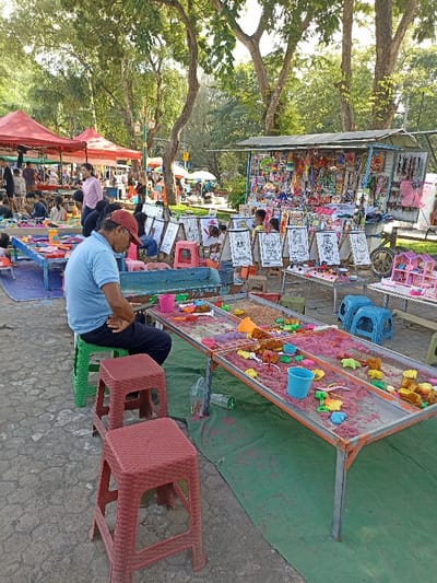 The image depicts a lively outdoor scene, likely a recreational park or market area in Mataram, Indonesia, under bright sunlight. In the foreground, a man wearing a blue polo shirt and a red baseball cap is seated on a green plastic stool, appearing to be resting or observing, with his hands clasped. Two empty red plastic stools are positioned near him.
Adjacent to the man is a long, low table offering a kinetic sand play activity. The table features multiple compartments filled with both natural and pink-colored sand, along with numerous colorful plastic sand toys such as molds, shovels, and buckets. A green tarp is laid out underneath this table.
In the midground, several children are actively engaged in coloring at individual white easels, which display various line drawings of cartoon characters, including a recognizable Mickey Mouse and other animated figures. These children are seated on small, brightly colored stools. Further back and to the left, under red canopies, other stalls or activity areas are visible with more people.
On the right side of the frame, a vibrant stall is densely packed with a wide array of colorful merchandise, predominantly toys and small trinkets. A miniature dollhouse is also displayed on a separate smaller table in front of this stall. The background features lush, green trees, indicating a park setting. The overall atmosphere is busy and recreational, suggesting a popular spot for families and children. No prominent text is visible on signs or products.