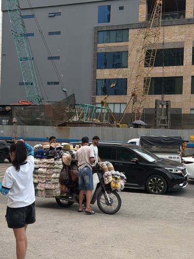 A group of people are gathered around a parked motorcycle laden with goods. The motorcycle is positioned on a paved surface, possibly a street or market. The individuals appear to be engaged in some form of transaction or preparation for sale. The motorcycle is heavily loaded with what appear to be rolled textiles or mats, some of which are bundled with string or plastic. The people are casually dressed, suggesting a warm climate. One person is standing with their back to the camera, wearing a white t-shirt and dark shorts. Another person, wearing a checkered shirt and denim shorts, is sitting on the motorcycle. Two other individuals are also visible in the vicinity, one standing and another partially obscured.
In the background, a large building with a textured facade and numerous windows is visible. A construction crane is also present, indicating ongoing development or building work in the area. Another vehicle, a black car, is parked next to the motorcycle. The scene appears to be taking place during the daytime, with ample sunlight casting shadows. The overall atmosphere seems bustling, characteristic of a commercial or urban environment. There is some visible text on the black car, which appears to be a license plate number. The context provided indicates the location is Đà Nẵng, Vietnam.