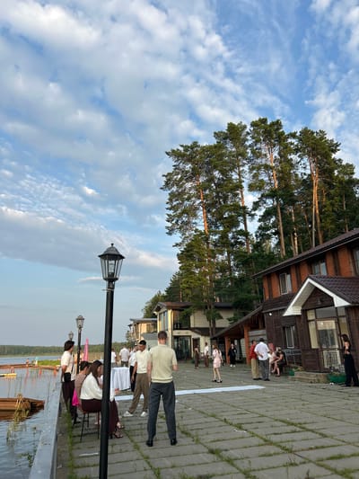 The image depicts an outdoor gathering on a paved walkway adjacent to a body of water, likely a lake or river, with a line of wooden structures and tall pine trees in the background. The sky is partly cloudy with a blend of blue and white. Several people are present, some standing and conversing, while others are seated. One group is gathered around a white-draped table. A tall, black lamppost is visible in the foreground. The overall atmosphere suggests a social event or celebration. The location is identified as the city of Chaykovsky, Russia.