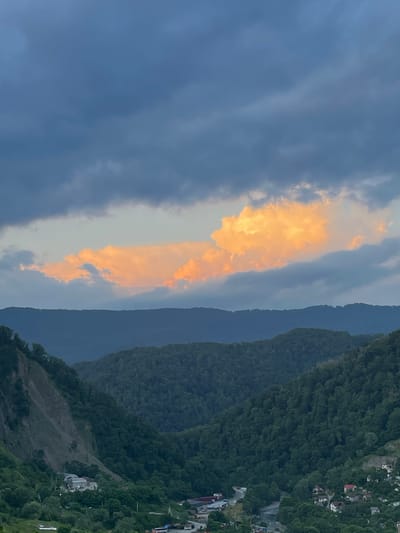 The image shows a mountainous landscape in Baranovka, Russia, with a dramatic sky. The scene is dominated by rolling hills covered in dense, dark green forests. In the foreground, there are visible signs of human habitation, with small buildings and structures nestled into the slopes of the mountains. A road or path winds its way through the terrain, connecting the houses.

The sky is a striking feature, with a large swath of bright orange and yellow clouds cutting through a more somber, grey-blue sky. This contrast suggests either sunrise or sunset, with the warm light illuminating the clouds. The weather appears to be changing, with the darker clouds hinting at potential rain or a shift in atmospheric conditions.

The overall mood is one of natural beauty and perhaps a touch of dramatic intensity due to the sky. The location cues of dense forests, mountainous terrain, and scattered settlements are consistent with a rural or semi-rural area in Russia. No people or specific objects are clearly discernible as primary subjects, the focus being on the interplay of light, weather, and the natural environment. There is no visible text in the image.