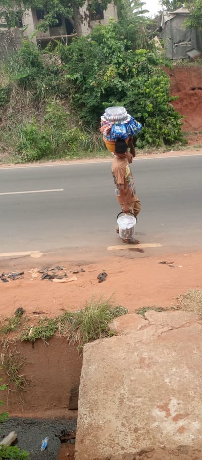 A young male, identified as a street vendor, stands on a reddish-brown dirt roadside shoulder adjacent to a two-lane asphalt road with a solid white edge line, in Ogidi, Nigeria. He wears a dark beanie, a light brown t-shirt with a graphic print, light-colored shorts, and open-toed sandals. He carries a large, multi-tiered display of packaged snacks and candies, including a prominent silver container on top, balanced on his head. In his left hand, he holds a clear plastic bucket containing numerous dark-colored items. The dirt shoulder exhibits scattered black plastic refuse. Behind the vendor and the road, dense green vegetation, including bushes and trees, covers an upward-sloping embankment. In the background, a partially constructed concrete building is visible through the trees on the left, and a tarp-covered structure with associated metal debris is seen on the right. The lower foreground reveals a concrete slab and a drainage culvert containing dark fluid and a blue plastic wrapper.