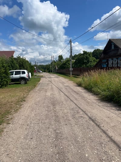 A dirt road winds through a rural area in Vyazniki, Russia, flanked by dense green foliage and wooden structures. On the left, a white SUV is parked amidst tall evergreen bushes. Further down the road, a person can be seen near a wooden fence. To the right, a weathered, dark wooden house with blue window frames stands out, bordered by tall, overgrown grass. Power lines stretch across the blue sky, which is dotted with white, fluffy clouds. The road itself is a mix of dirt and gravel, with tire tracks visible.
