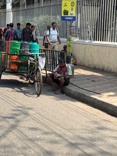 The image depicts a street scene in Kolkata, India. A group of people are walking alongside a road. A cycle rickshaw loaded with colorful containers stands near the sidewalk. A man reclines on the sidewalk, near the curb and a metal guardrail. A sign promoting "Eurogrip" is visible on a post, along with other posters. The environment appears sunny, with shadows cast on the pavement.