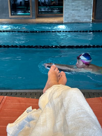 The image depicts an indoor swimming pool scene in London, UK, capturing both activity and relaxation. In the central and left portions of the frame, a person is actively swimming laps. They are wearing a distinctive blue, white, and pink swim cap and goggles, with their head partially submerged in the clear blue water as they execute a swimming stroke, creating visible ripples. Black lane lines segment the pool along the swimmer's left side.
In the foreground, on the right, the bare feet and lower legs of another individual are visible, resting comfortably on a reddish-brown, textured poolside surface. A soft, white towel is draped over their legs, partially obscuring them. A tag on the towel is clearly visible, bearing the text "100% COTTON," alongside other smaller, indistinct words.
The background shows reflections in framed elements, possibly mirrors or windows, on a wall adjacent to the pool, highlighting the indoor setting. The lighting suggests an artificial indoor environment, indicative of a time when natural light is not dominant, such as evening. The scene portrays a balance between focused physical activity and serene poolside observation or rest.