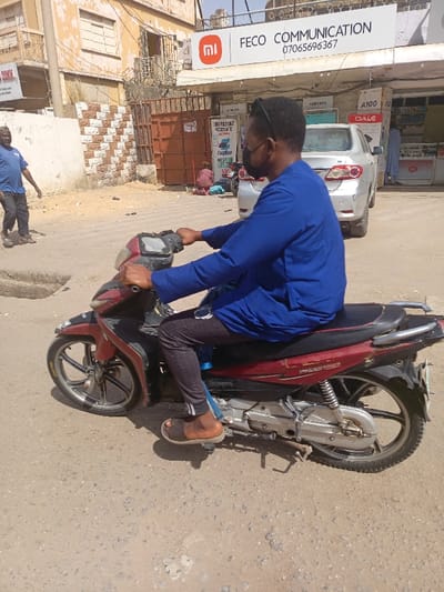 The image presents a street scene in Fagge A, Nigeria, under bright daylight. In the foreground, a man with dark skin is seated on a red and black motorcycle, holding the handlebars. He wears a blue long-sleeved tunic, dark patterned trousers, brown slides, a black face mask, and sunglasses perched on his head. The motorcycle is identified as a 'PASSPORT' model, featuring silver-colored engine components and spoked wheels.
To the immediate left of the central figure, a second man, wearing a blue t-shirt and dark pants, walks towards the left, with his left foot raised. Behind him, a light-colored building with a section of brown and white checkered wall is visible, alongside a rusted metal gate topped with barbed wire.
In the background, a commercial storefront is present. A prominent white sign above it displays a red 'Mi' logo, the text "FECO COMMUNICATION," and a phone number "0706569367." The storefront features product displays, including signs for "SAMSUNG," "A100," and "OALE." A silver sedan is parked in front of this store, partially obscuring the entrance. Near the storefront entrance, a person dressed in red/pink clothing is seated on the ground. Another sign advertising "COMPUTERS SERVICE" and "EasyBuy" is also visible.
The ground surface is an uneven, light-colored, dusty asphalt or dirt road.