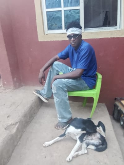 A man sits on a green plastic chair on a concrete porch in front of a reddish-brown building. He wears sunglasses, a white headband, a blue t-shirt, and ripped light-blue jeans. His feet are clad in sandals and dark sneakers. A window is visible behind him. A black-and-white dog lies on the concrete, licking itself near the man's feet. A dark object, possibly a suitcase, sits to the right of the chair.