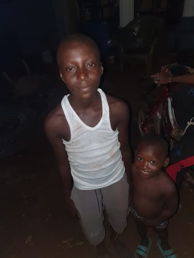 This dimly lit vertical image, possibly taken at night in Idah, Nigeria, features two young boys looking towards the camera and smiling.
In the foreground, a boy is positioned towards the left side of the frame. He has dark skin and a short haircut, looking directly at the camera with a gentle smile. He is wearing a white sleeveless tank top and light grey pants with some faint, possibly Arabic, text visible on the left leg, appearing like "لل".
Behind him, slightly to the right and appearing to be at a higher elevation, is a second boy. He also has dark skin and a broad smile, looking playfully towards the camera. This boy is bare-chested and wears patterned shorts or underwear. Light blue sandals are visible on his feet. An "adidas" logo can be seen on a slipper worn by an adult hand behind him.
Partially visible behind the second boy and the adult hand is the red and white frame of what appears to be a motorcycle or scooter. The adult's hand, adorned with a bracelet, rests on a handle of this vehicle. The background is mostly dark and indistinct, suggesting an indoor setting or a very poorly lit outdoor area. The overall mood of the image is joyful and candid, captured with direct flash in a low-light environment.